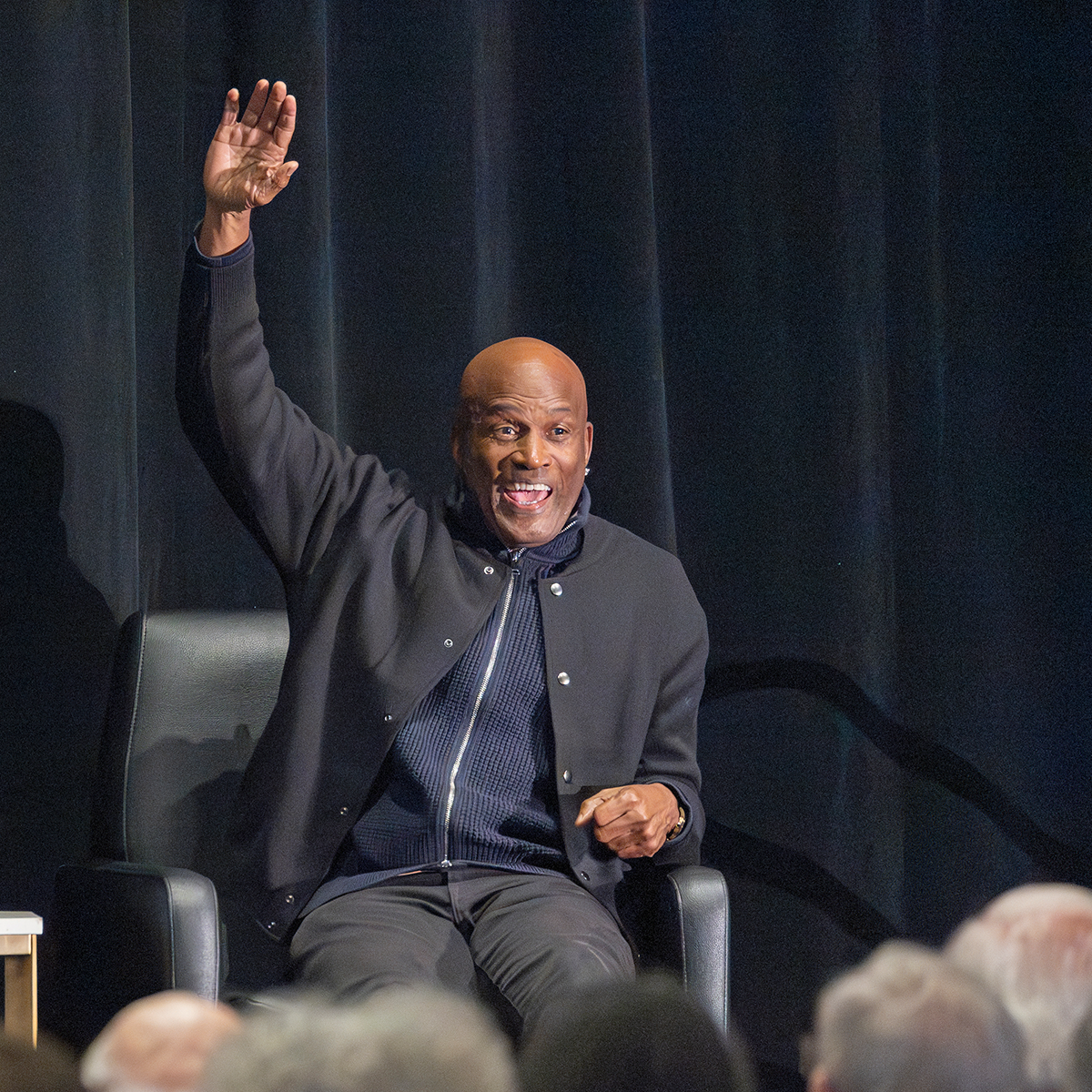 A seated person on stage raises their hand and smiles.