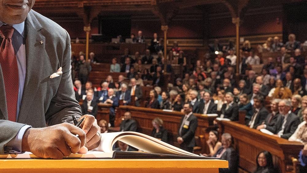 A person, wearing a business suit and tie, is writing in a book with white pages. He is holding a pen in each hand. We see part of his face, from his lips down. In the background is an audience seated in a large auditorium.