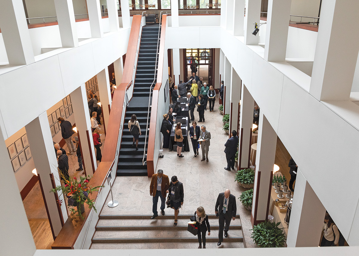 A wide shot of the lobby of the House of the American Academy of Arts and Sciences, an atrium that joins a two-story space with two sets of stairs. Natural light fills the space and dozens of people walk throughout.