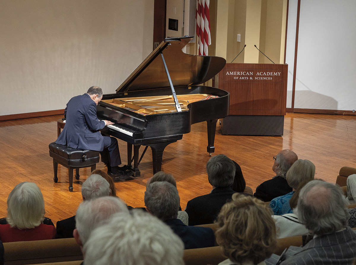 An audience watches a man dressed in a blue business suit play a piano in a well-lit auditorium.