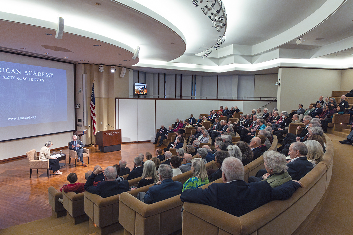 An auditorium full of people watching two speakers on stage in conversation.  