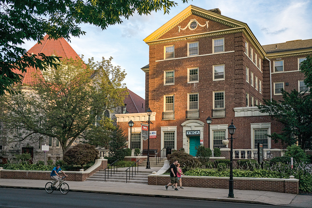 People stroll on the sidewalk and bike in the street outside of a YWCA at dusk.