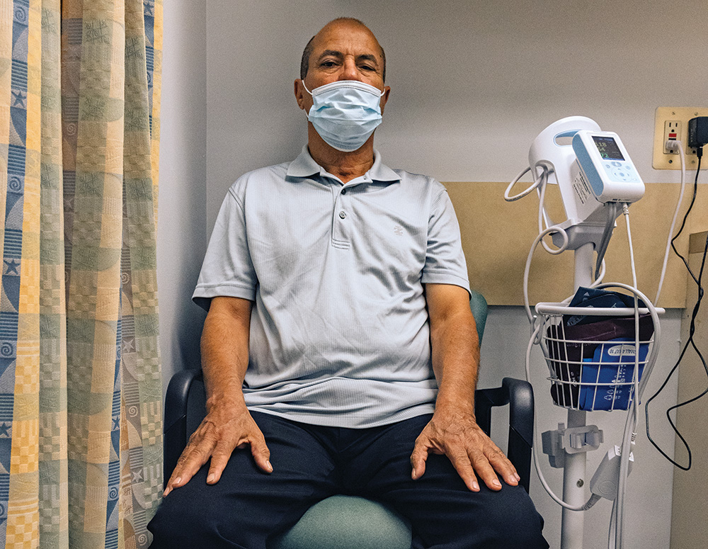 A person with dark skin wears a face mask and sits in a chair beside triage equipment, looking steadily at the viewer.