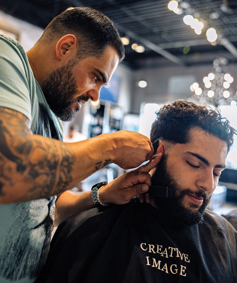A barber applies an electric razor to the hair behind the ear of a customer. 