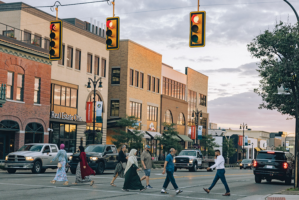 People pass each other in a crosswalk in a prosperous shopping area. Cars idle in the street.