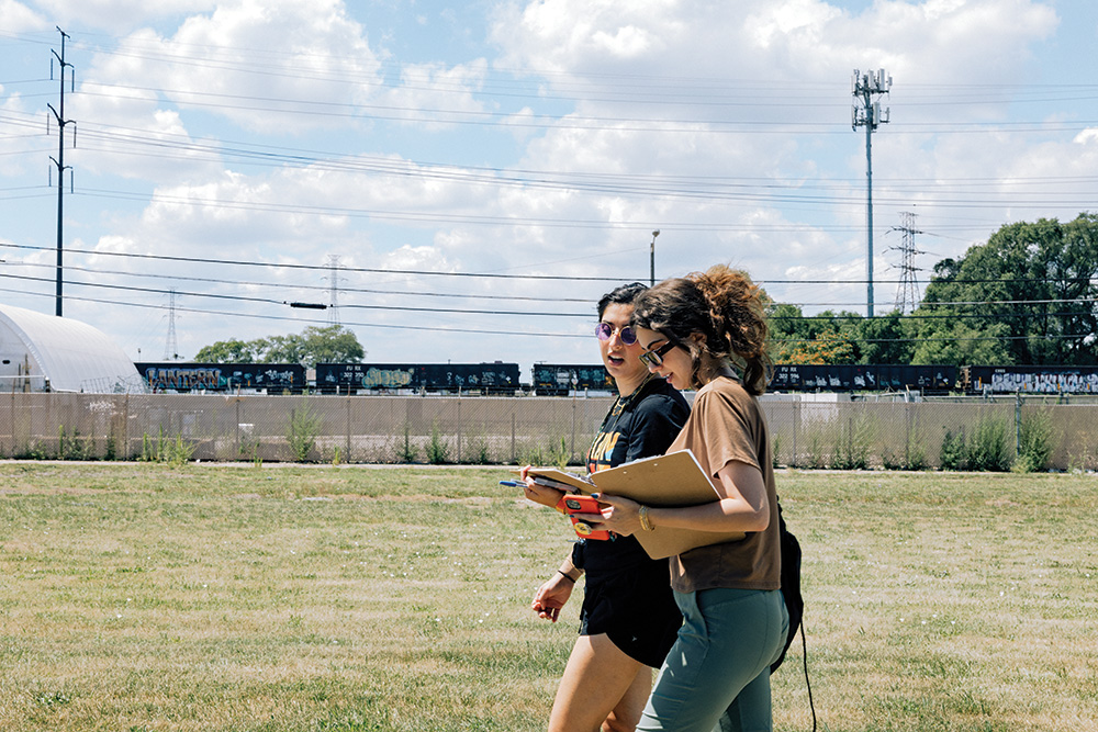 Two people walking while holding clipboards. A train runs in the background. 