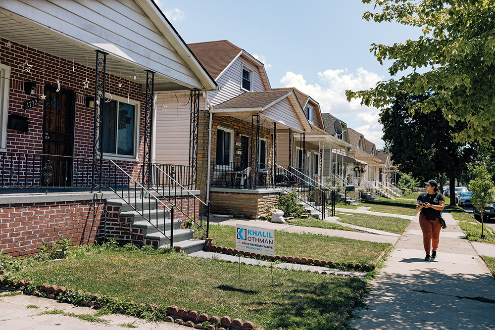 A person with brown skin walking along a sidewalk holding a clipboard. One of the houses has a political candidate sign in the yard. 