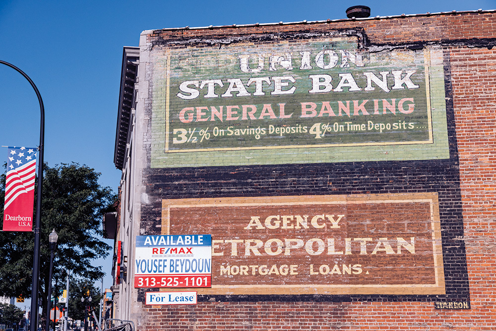 A red brick wall with worn advertisements for “Union State Bank: General Banking: 3½% on Savings Deposits 4% On Time Deposits…” and “Agency Metropolitan: Mortgage Loans.” A “For Lease” sign with contact information for a real estate agent obscures the “M” in “Metropolitan.”