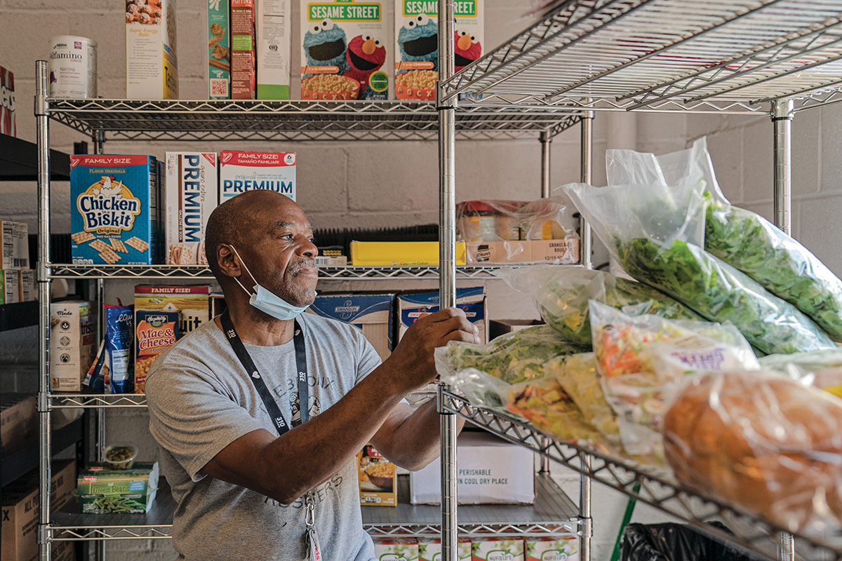 A man with brown skin staring into the distance while he grabs onto a metal shelf full of uncooked food. There is another shelf behind him filled with packaged groceries.