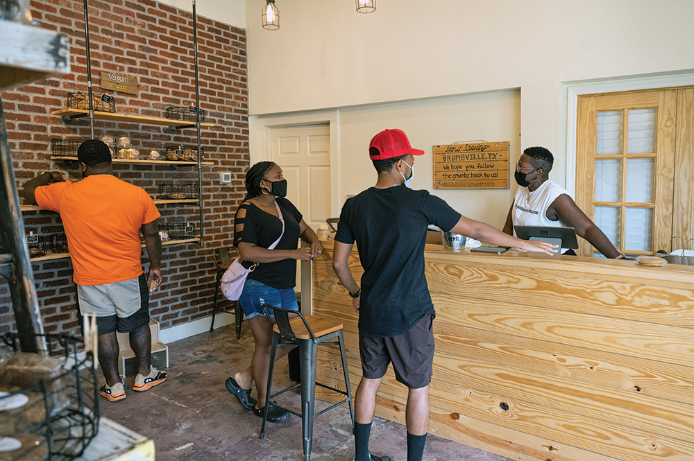 People with brown skin wearing face masks shopping at a bakery. The owner stands behind the counter. 