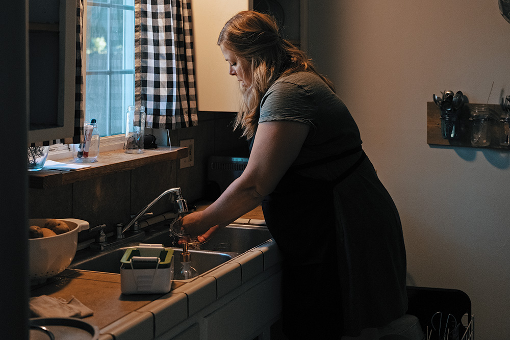 A person with pale skin washing dishes in their home in a darkly lit kitchen. 