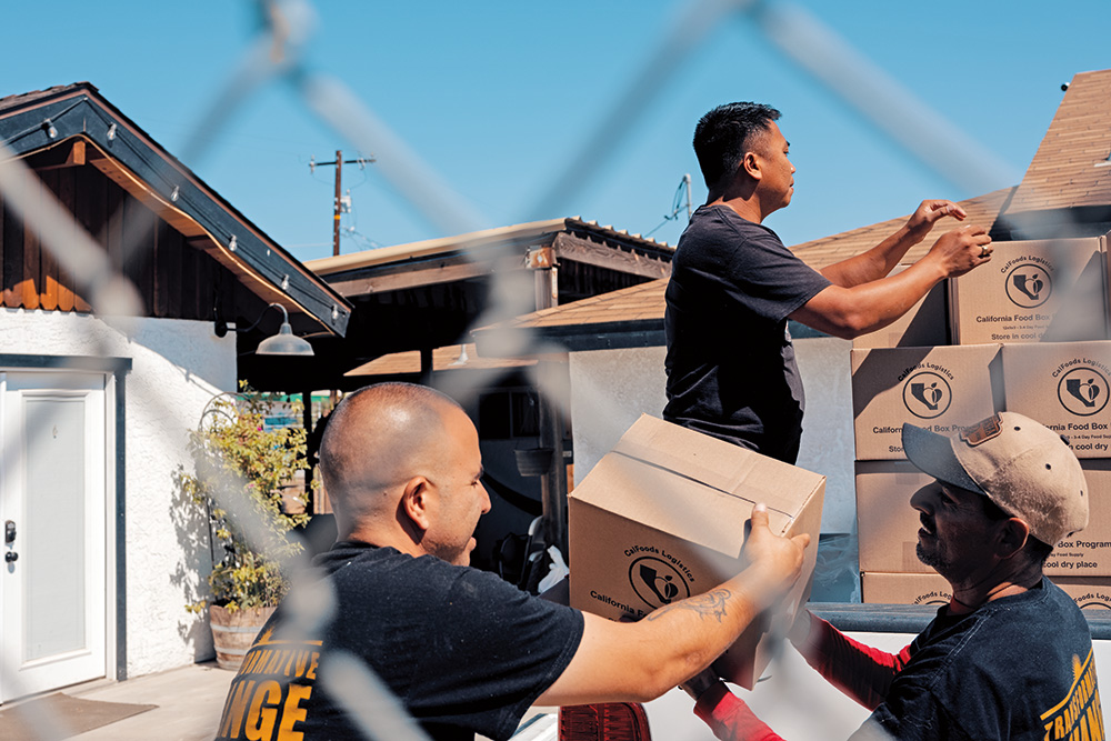 Three people load boxes onto a truck. Two are holding one box, while another person is reaching for the next box. 
