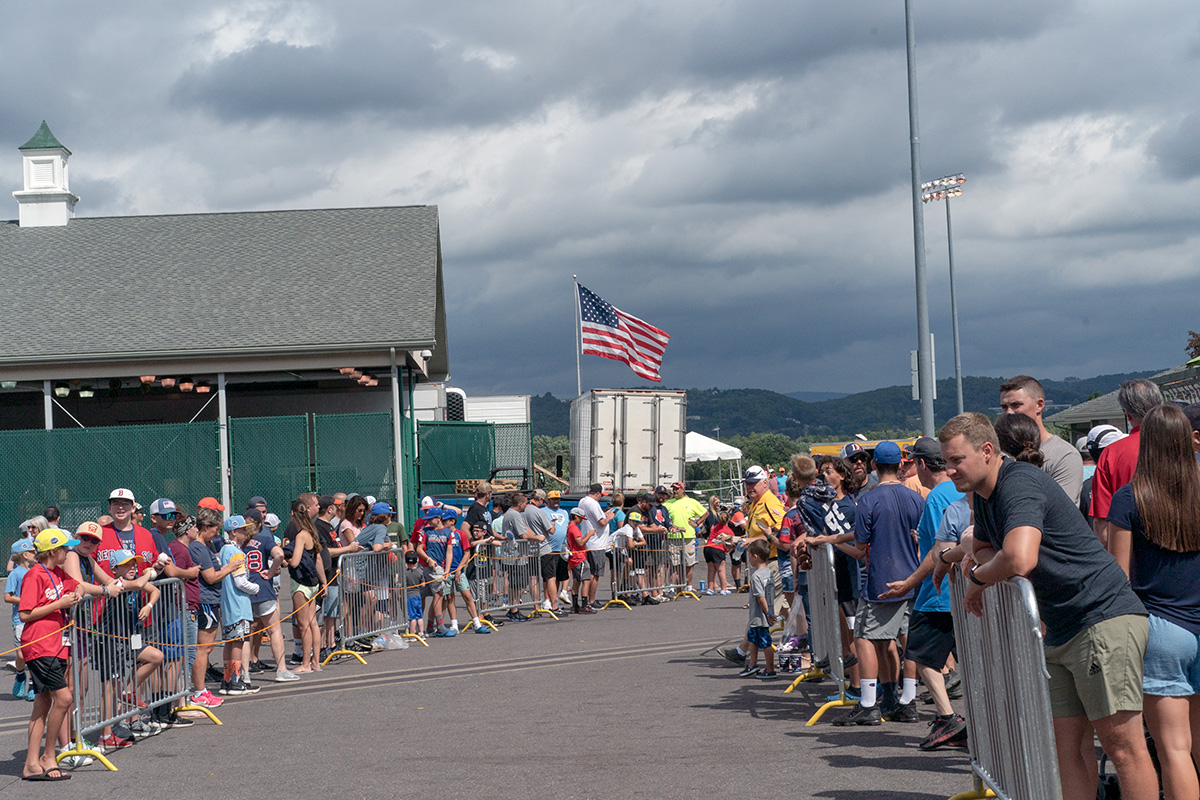 People lining a fence around a cleared walking area. An American flag flies in the background. 