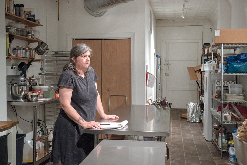 A person with pale skin with their hands on a steel table in the backroom of a tea shop. Their face indicates concern. 