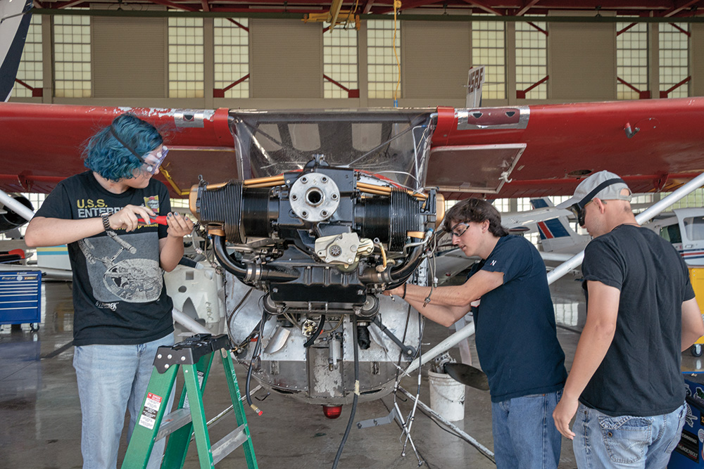 Three people who are wearing protective goggles work on the front of an airplane that is red and white. Two are holding tools while one is looking at the plane. 