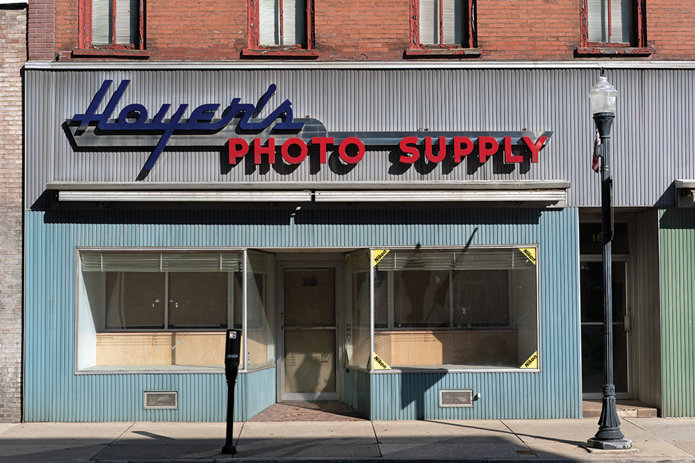 A vacant storefront with empty windows. Stickers in its right window announce Nikon cameras were once sold inside.