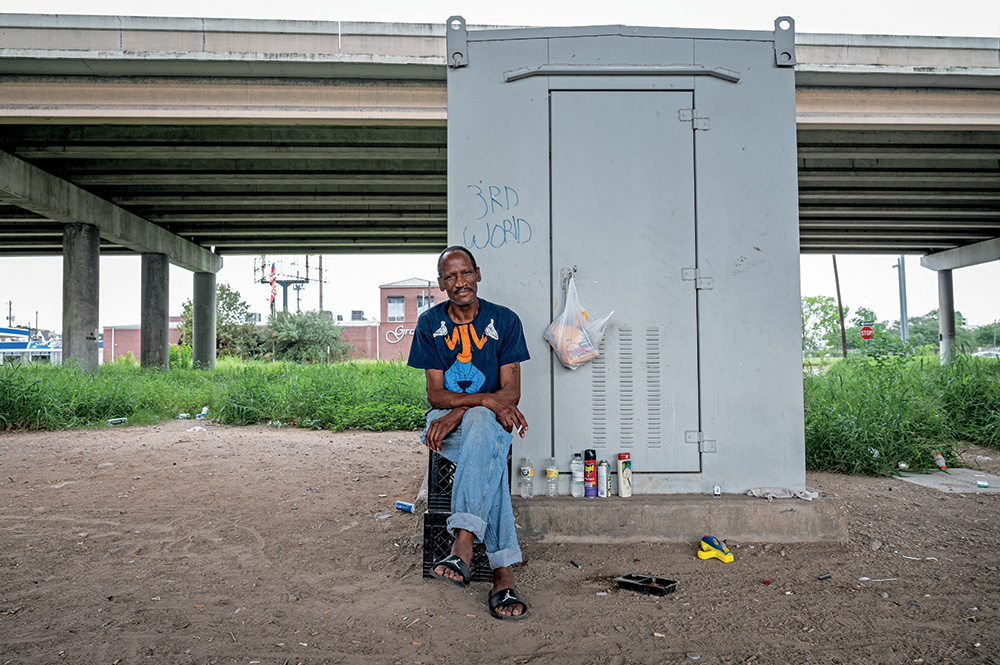A person with brown skin staring at the viewer. They are sitting on two milk crates in front of a power station under an overpass. The power station has “3rd World” written on it. 