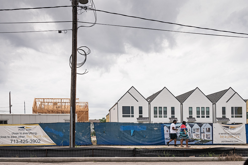 Two people walk in front of a series of new row homes. One home is under construction and four homes are completed. A sign advertising the new homes is attached to a fence in front of the buildings.