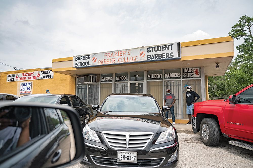 Outside of a barber training school. Two individuals in masks stand outside the school. The photographer’s camera is visible, reflected in their car’s side mirror. 