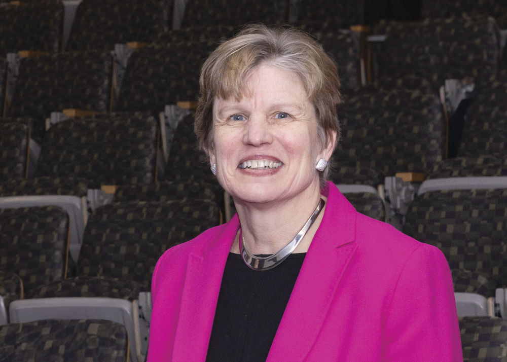 A fair-skinned woman with gray and brown hair smiles at the viewer. She is wearing a pink blazer and black top.  