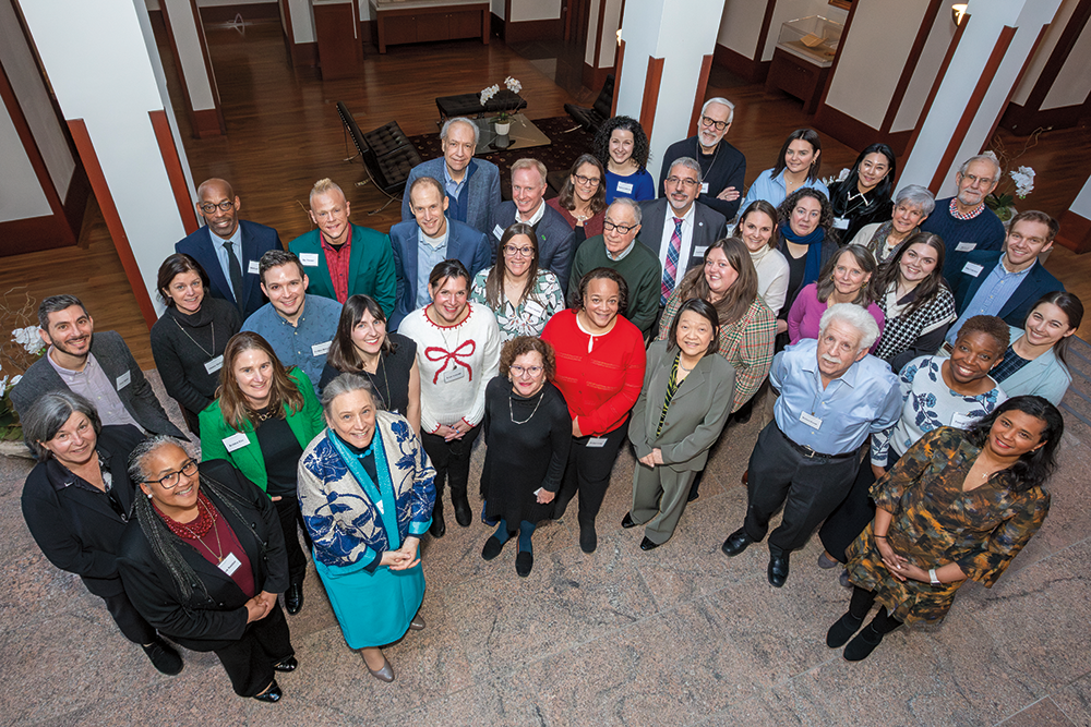 A group of 37 adults in business casual attire pose together for a photo taken from an elevated angle with everyone smiling up at the camera. The subjects in the photo are standing in a space with tiled flooring and white columns with wooden trim. 
