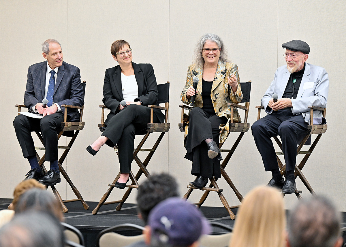 Four people, sitting in black director’s chairs, face an audience. The third person on the right is holding a microphone in their hand and speaking to the audience.  The people in director’s chairs to her left and right are smiling.