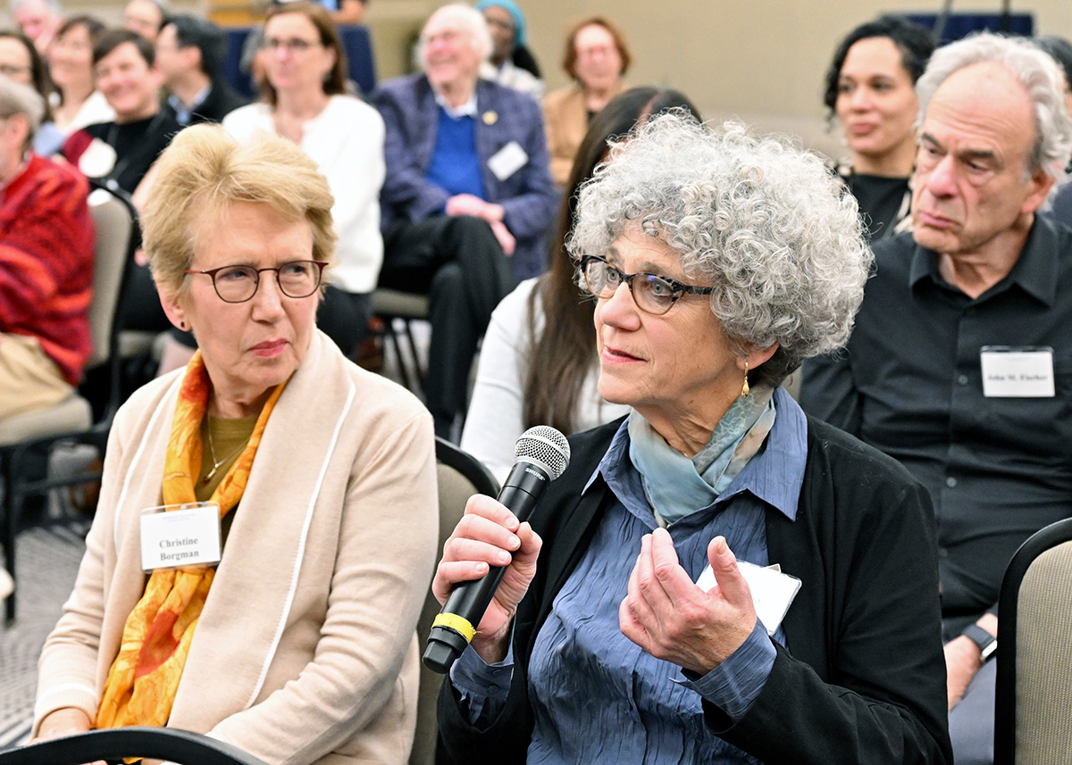 A light-skinned woman with curly gray hair, wearing dark glasses, a blue blouse and scarf and dark sweater and holding a microphone in her right hand, asks a question. She is seated in the audience, with several people around her.