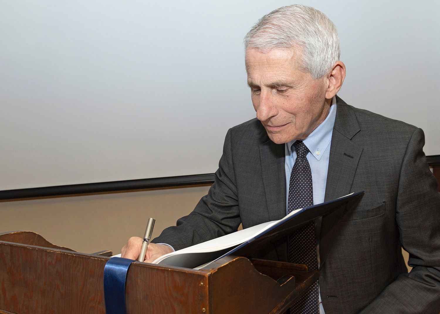 A photo of Dr. Anthony Fauci signing a page in a book that is open on a podium. He is looking down at the page. 