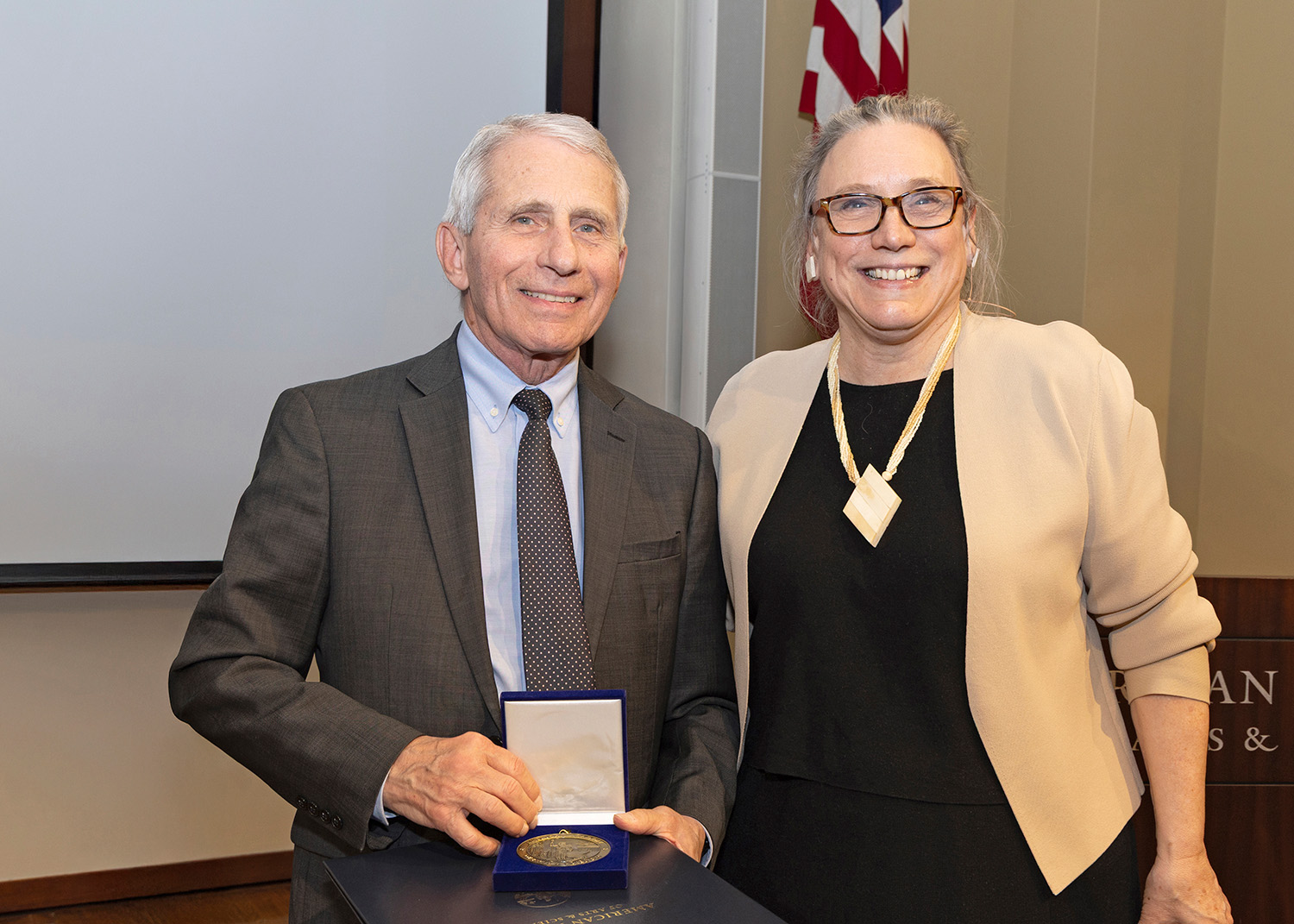 A photo of Dr. Anthony Fauci standing next to President Laurie Patton. Both are facing forward and are smiling at the viewer. Dr. Fauci holds an open case with a bronze medal inside. 