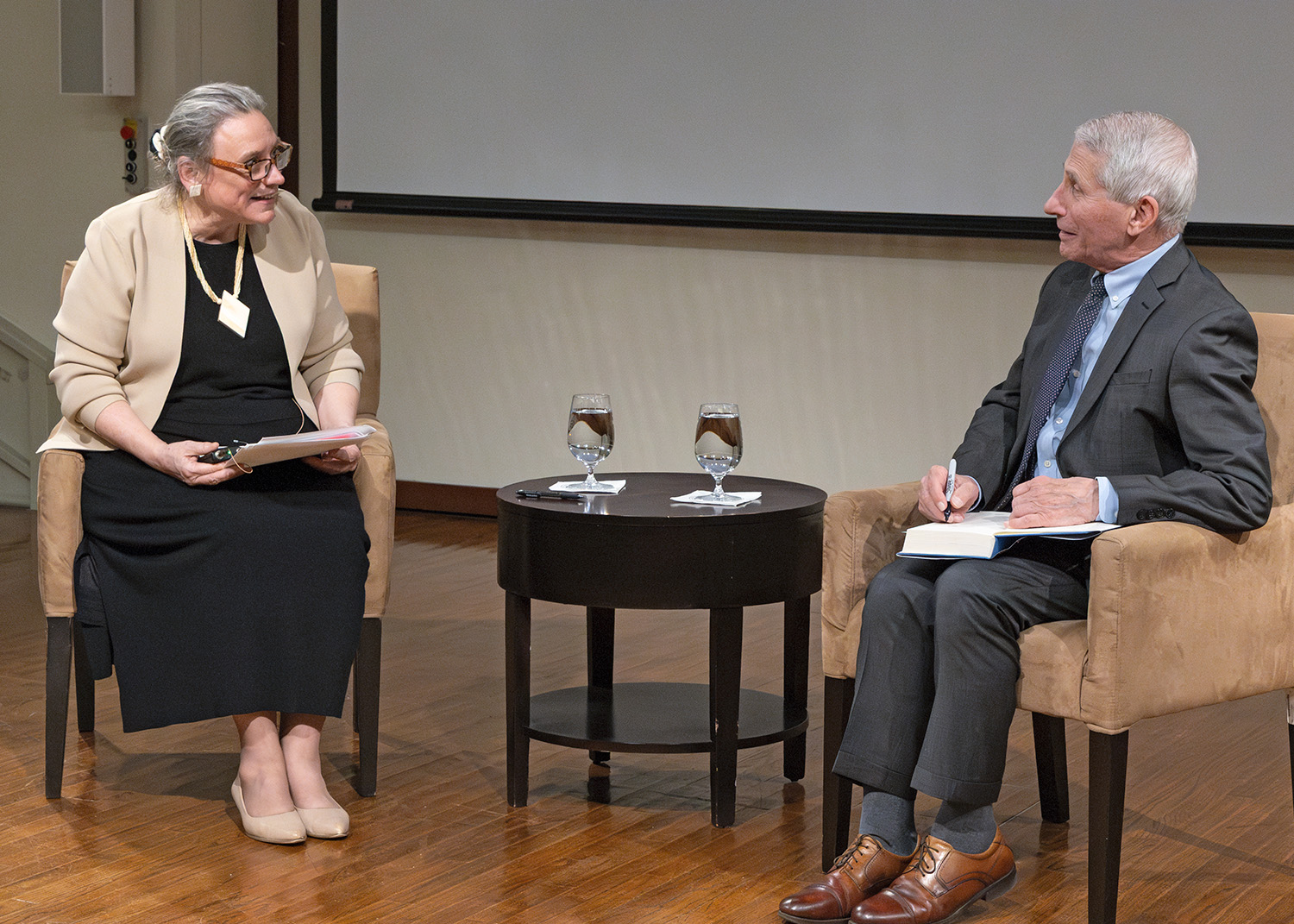 A photo of President Laurie Patton and Dr. Anthony Fauci, seated in chairs with a small round table between them. They are looking at each other. President Patton holds several papers in her hand. Dr. Fauci has an open book on his lap and holds a pen in his right hand. 