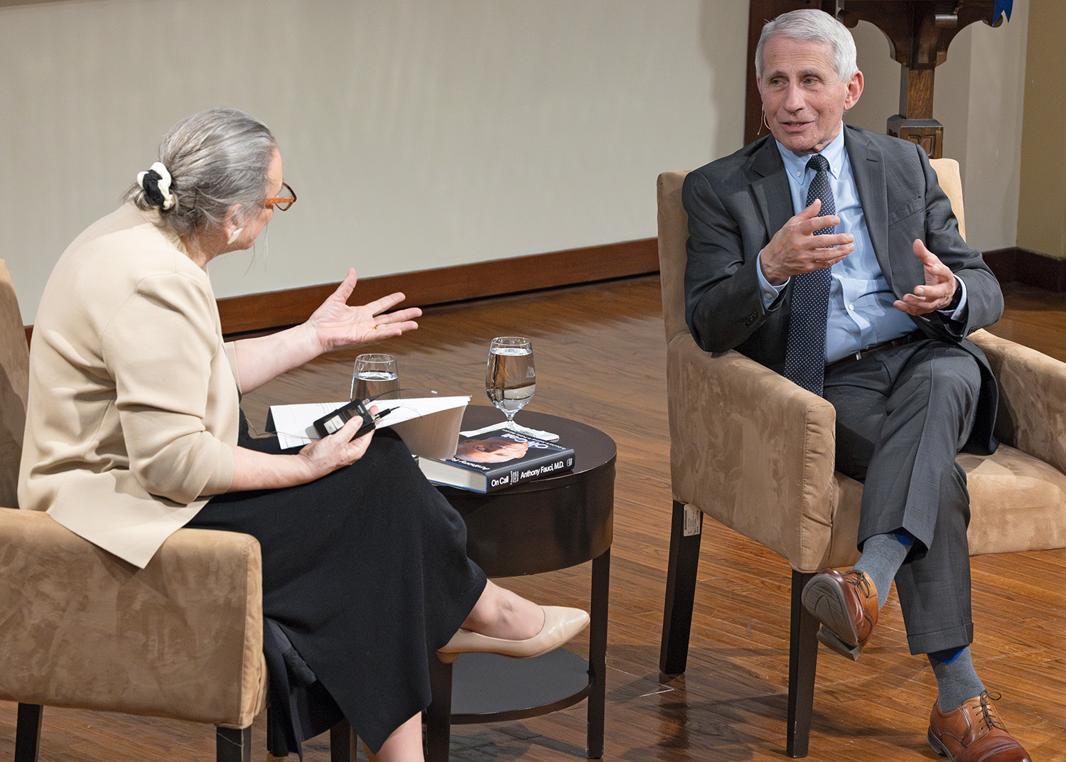 A photo of President Laurie Patton and Dr. Anthony Fauci in conversation. Both are gesturing with their hands.  