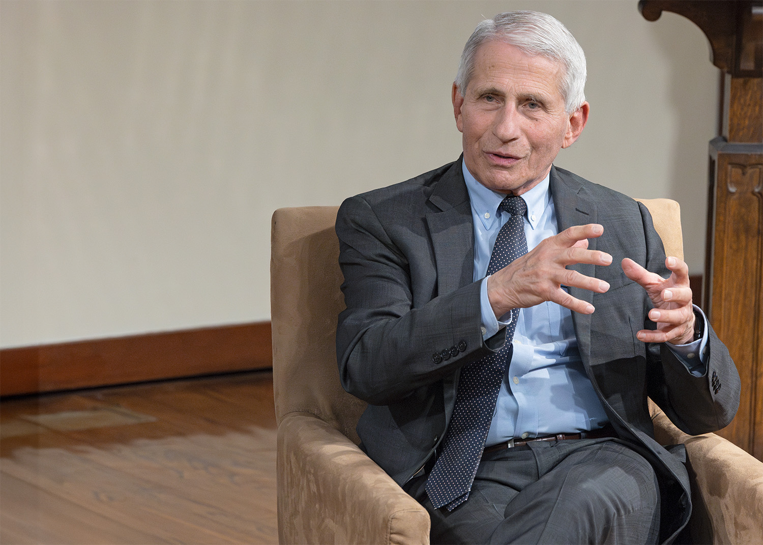 A photo of Dr. Anthony S. Fauci. He is seated in a chair and gestures with his hands as he speaks to an unseen audience. He has short gray hair and is wearing a dark business suit.  