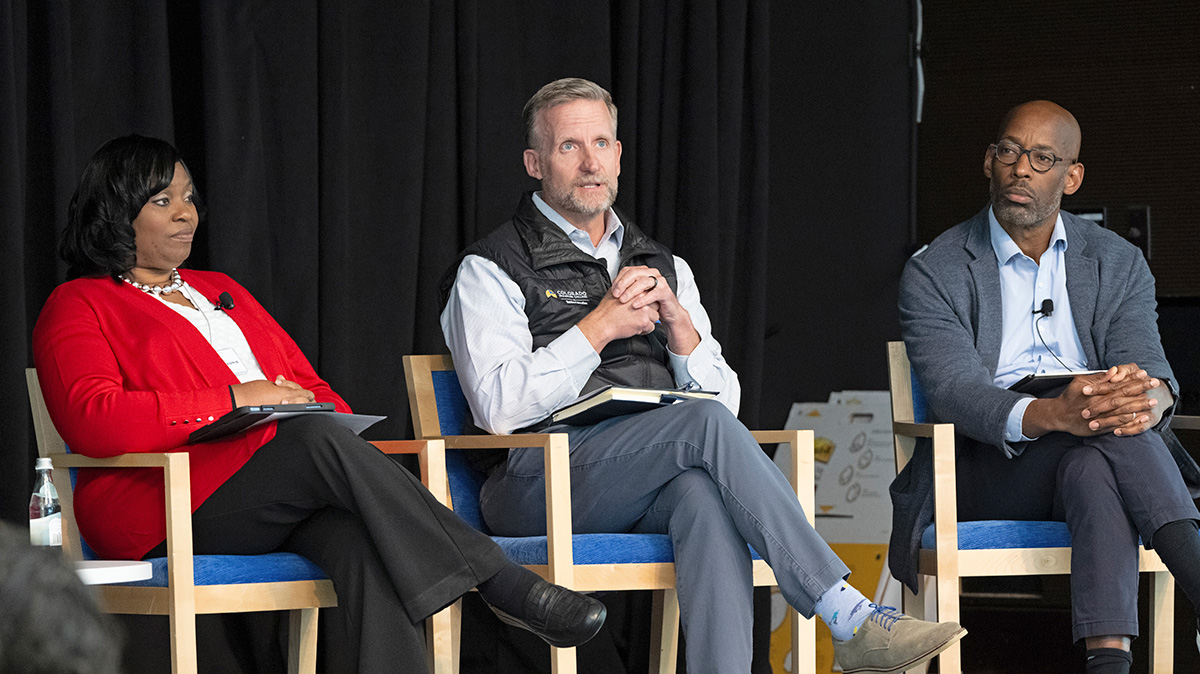 A woman and two men are sitting in chairs next to each other on a stage with a black background. The man in the middle is speaking and the other two individuals are listening.  