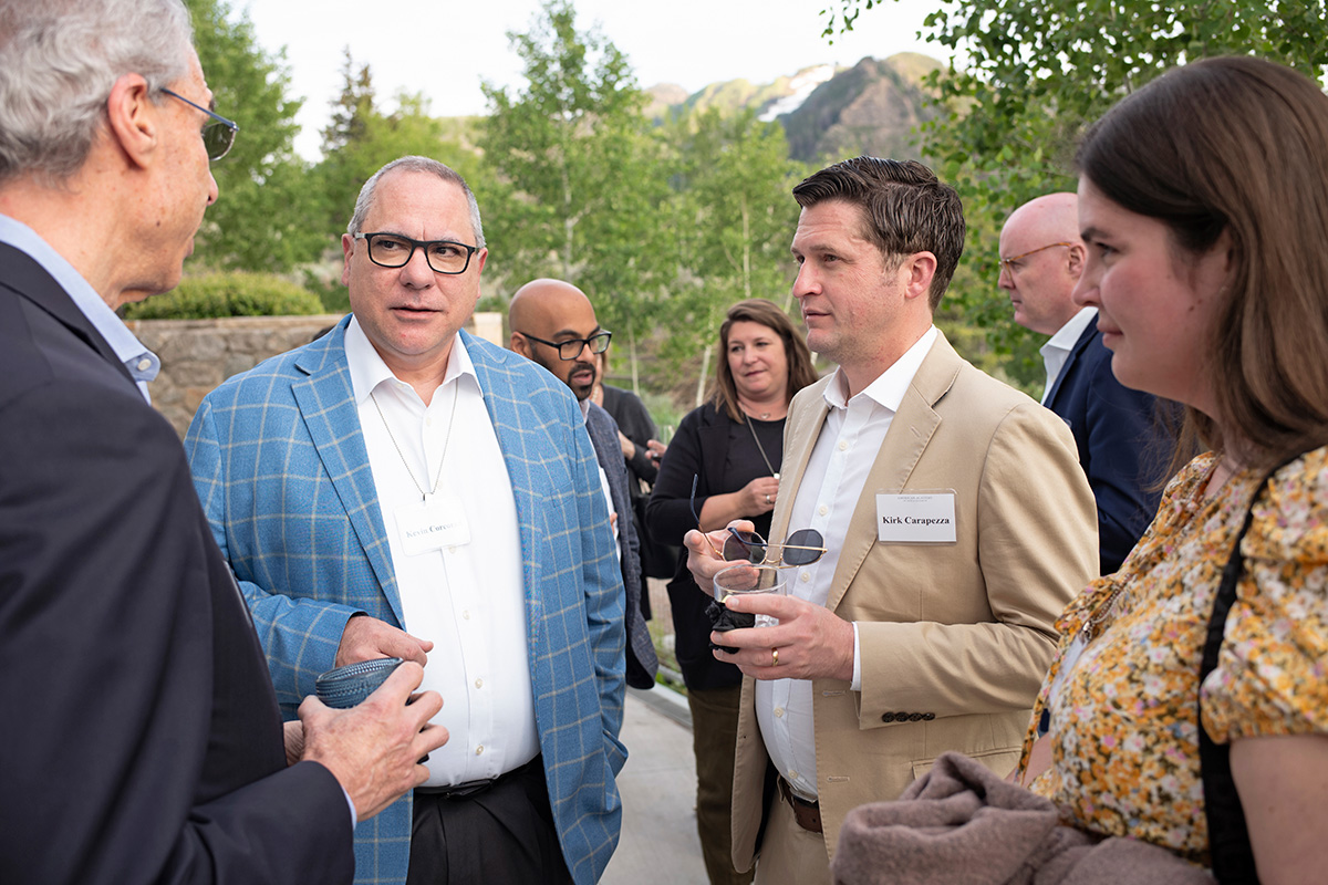 Four people are standing outside in a circle and talking. Trees and mountains are seen in the background. 