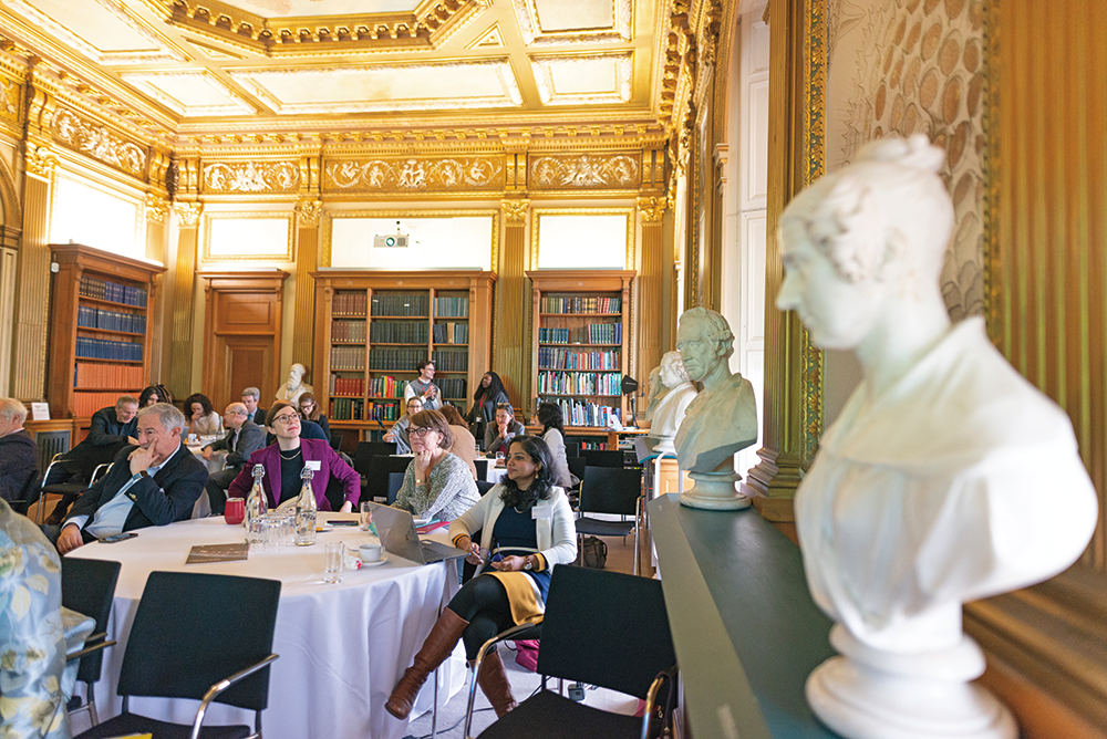 Groups of people sit at round tables draped in white linen in a room adorned with wooden bookcases and ornate gold trim. Several marble busts are placed around the perimeter of the room.
