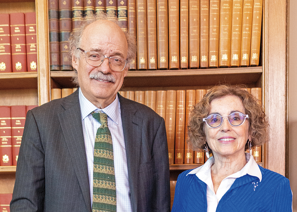 A photo of two people. The man on the left is wearing a charcoal suit and green and yellow tie. The woman on the right is wearing a blue suit and white blouse. They are standing in front of bookshelves and are facing the viewer.