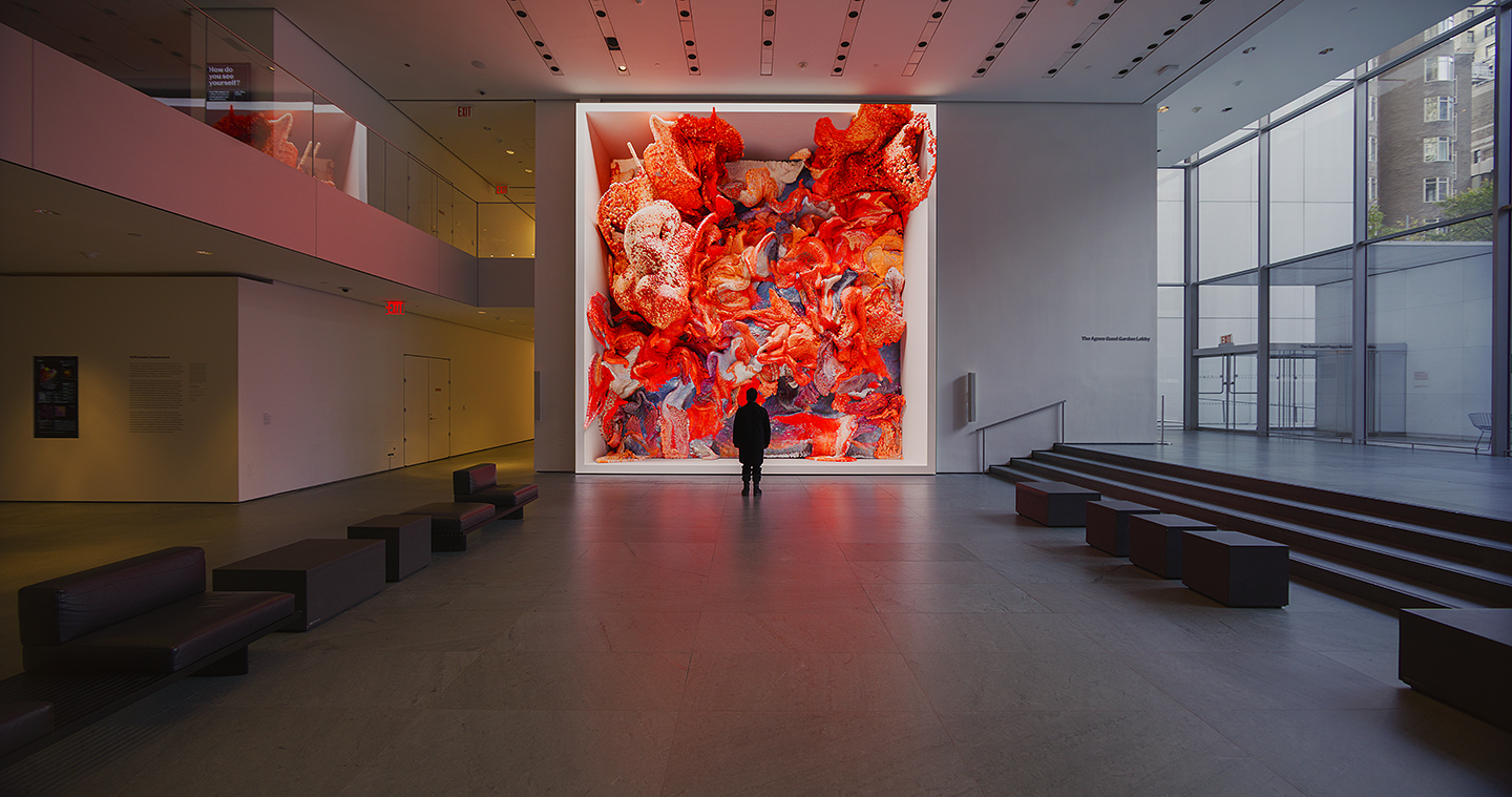 A person standing in front of a floor-to-ceiling screen in a large, open museum space. The image captures a frame of digital art in motion. The artwork resembles clouds of foamy material with bright orange-red and light gray colors.