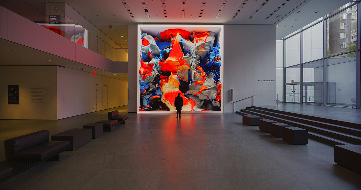 A person standing in front of a floor-to-ceiling screen in a large, open museum space. The image captures another frame from a work of digital art in motion, where the colors now include black and bright blue