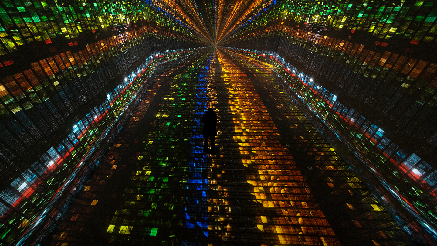 An overhead view of a person standing in a room displaying immersive digital art. The floor and walls are covered with small, multicolored squares (orange, green, blue, red). On the wall in front of the person, multiple rows of squares converge into a vanishing point.