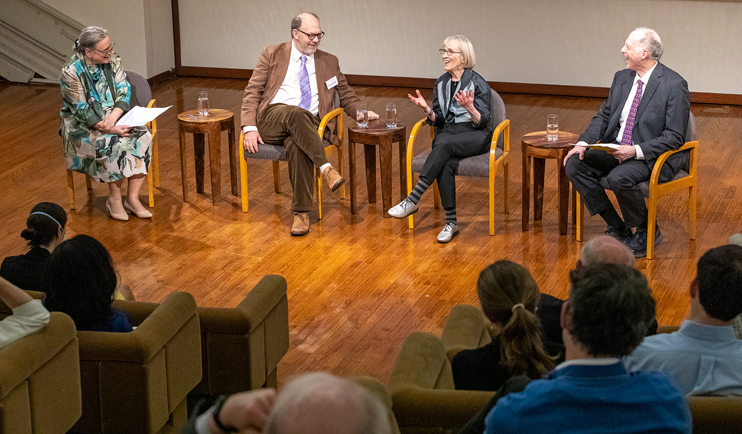 Four adults seated on wooden chairs on a stage are engaged in a discussion in front of an audience.