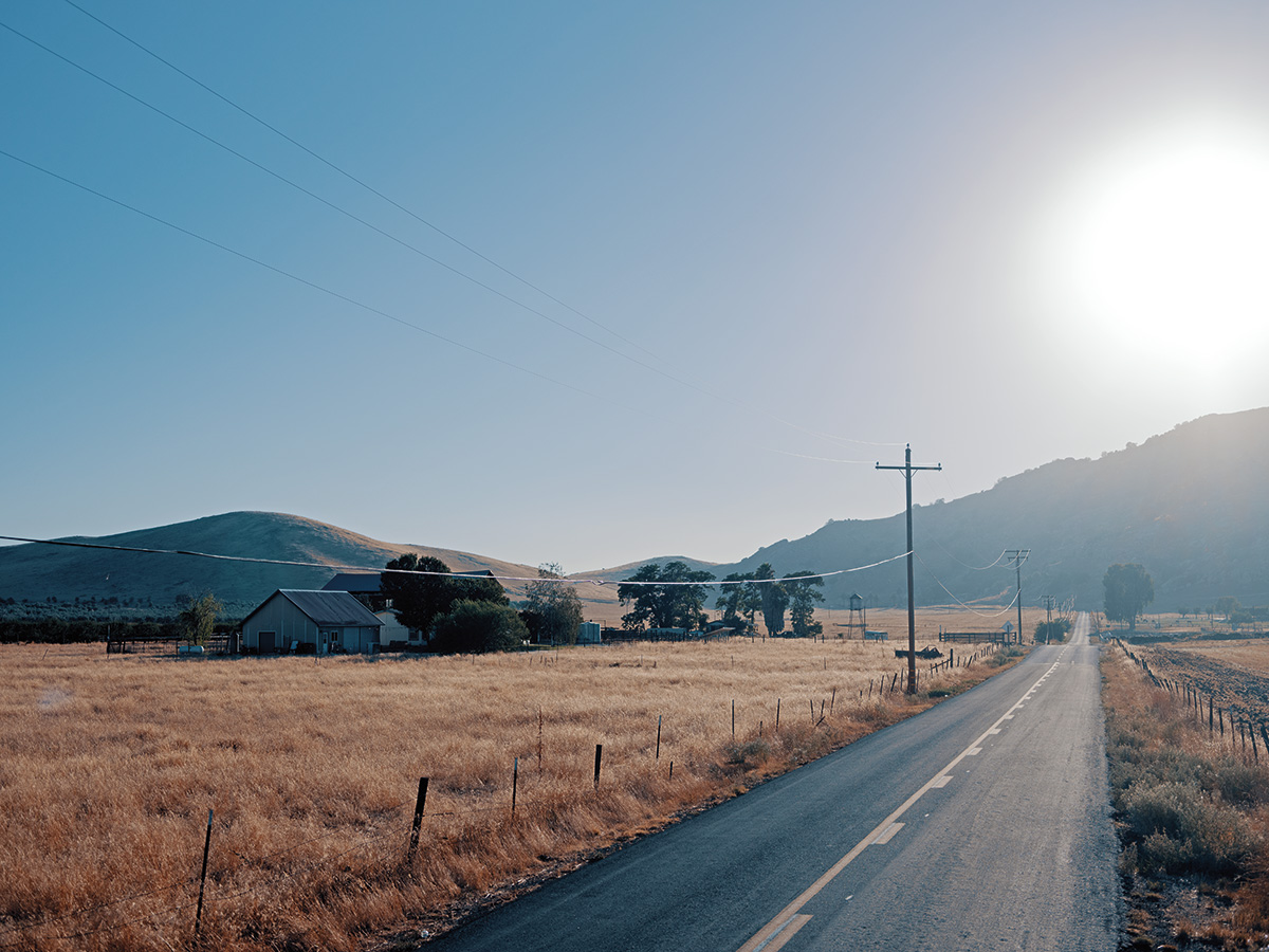 A photograph of a rural farm, divided by a road with powerlines on the left side and mountains in the background.