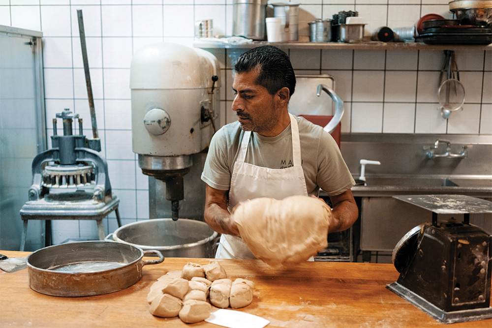 A person rolls dough in a bakery in front of a wooden counter. Dough that has already been rolled and cut sits on the counter. Pans and other bakery machinery are next to and behind them. 