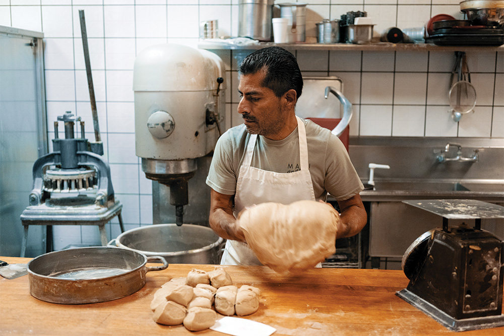 A person rolls dough in a bakery in front of a wooden counter. Dough that has already been rolled and cut sits on the counter. Pans and other bakery machinery are next to and behind them. 