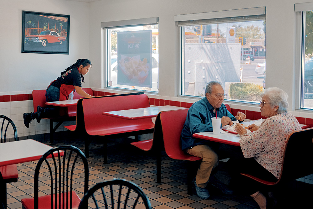 Two individuals eating a meal in a fast-food restaurant as a staff member wipes down a table nearby.