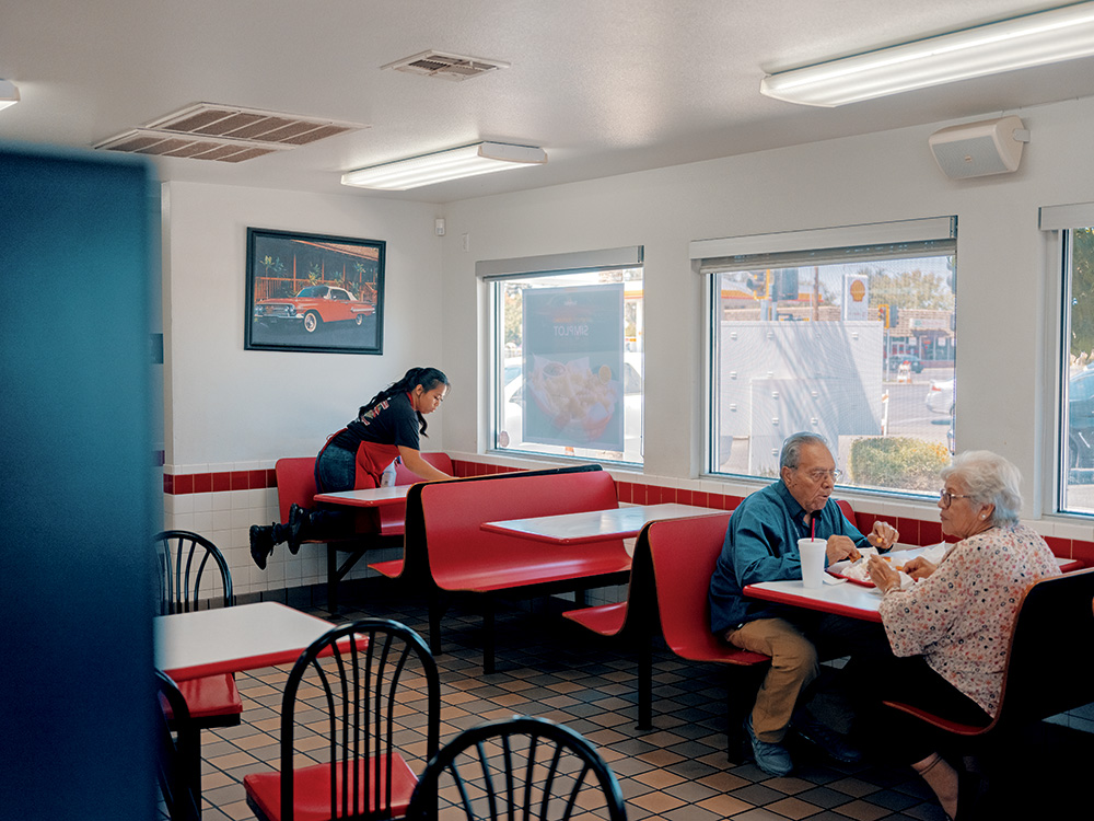 An employee in a fast-foot restaurant cleans a table. Ten feet away, a couple eats their food. The employee had long black hair and brown skin. The couple has white hair and gray hair, respectively, and brown skin. 