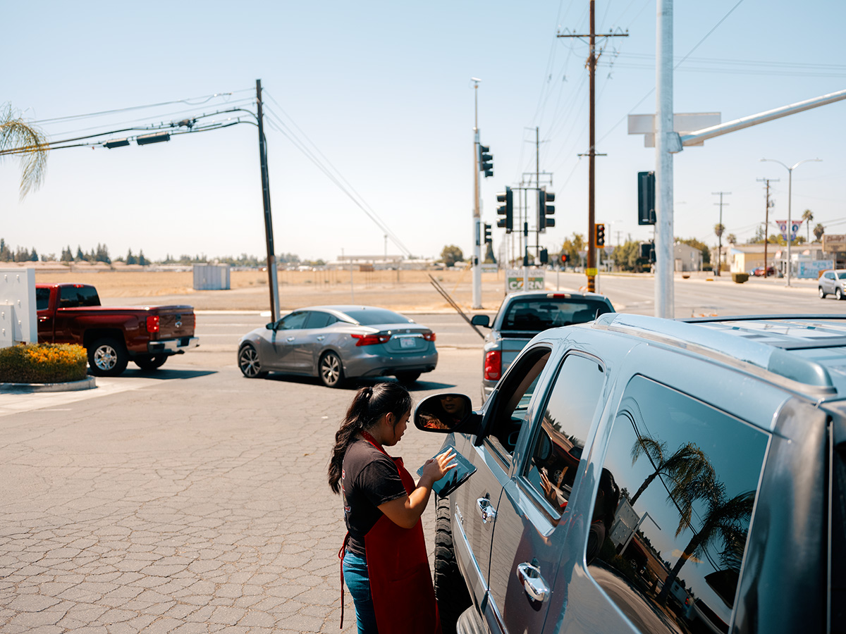 A fast-food worker taking an order using a table from a driver at the back of a line of cars. 