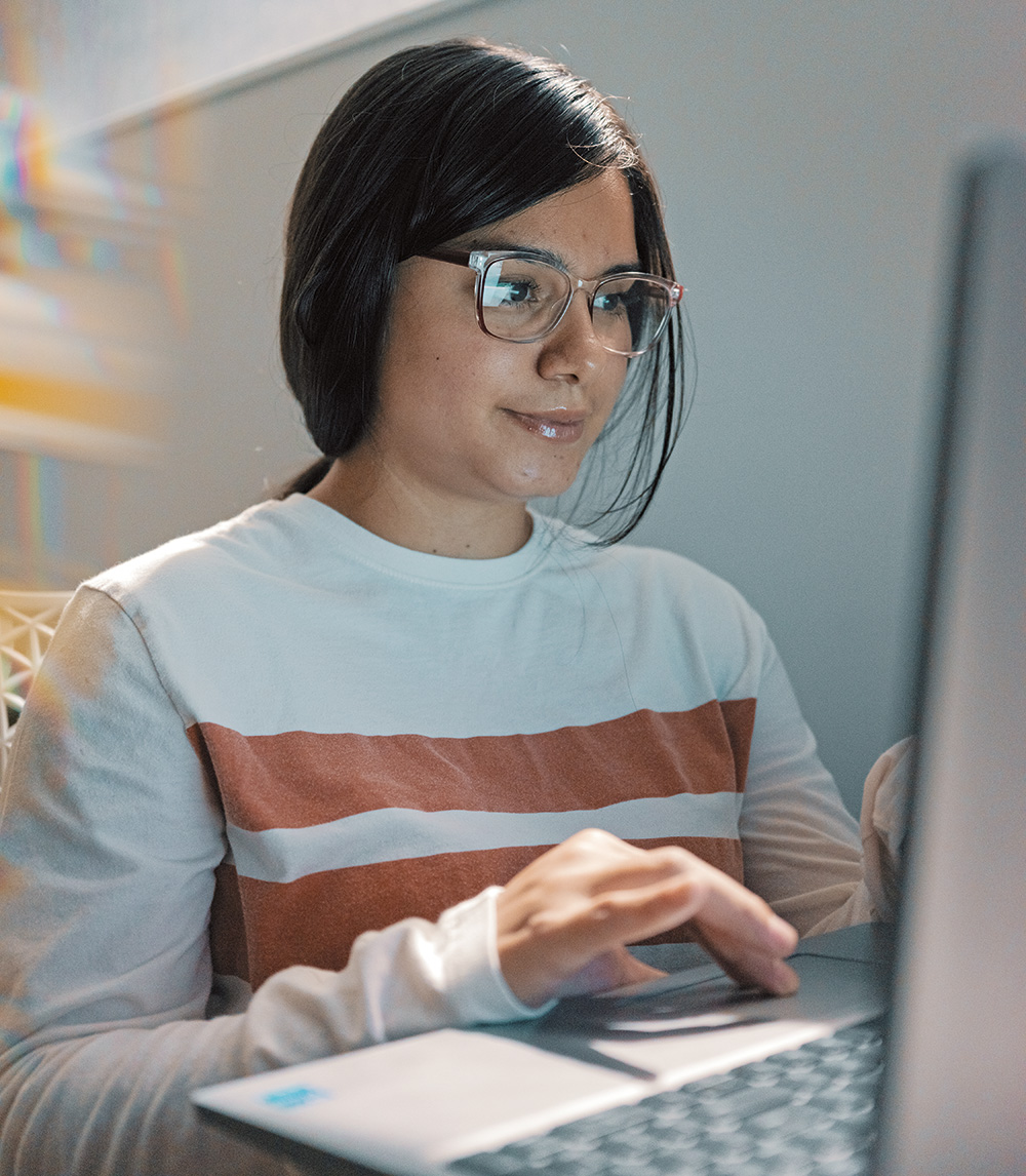 A person with their hair in a ponytail, wearing glasses, clicking on her laptop. 