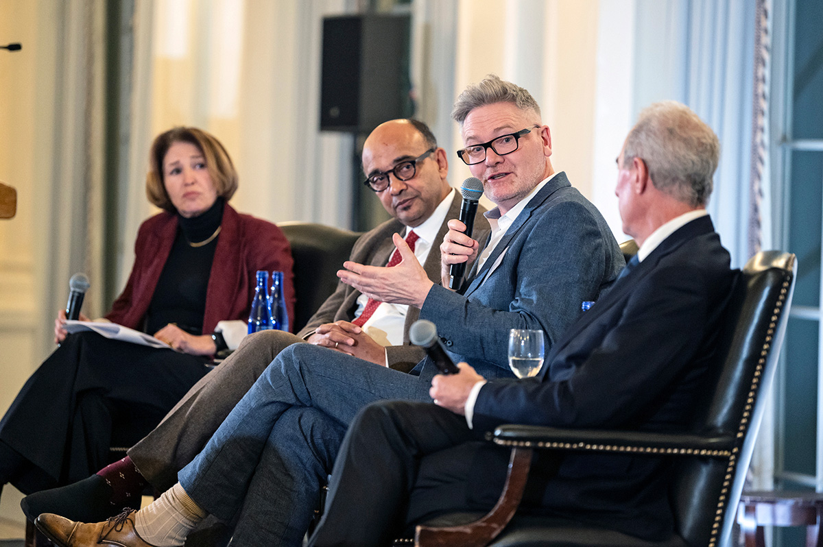 Four people are seated in chairs next to each other. The second person from the right is speaking into a microphone and looking at the person on their left.  The other three individuals are looking at the person who is speaking. 