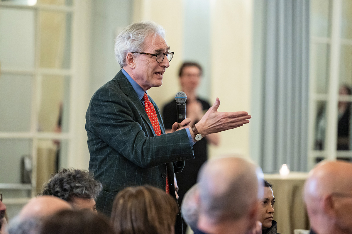 A light-skinned individual with short gray hair, wearing glasses, a red tie, and blue blazer, is standing and holding a microphone. He is addressing people who are not visible in the photograph. 