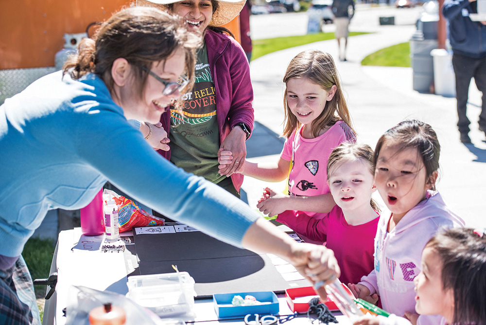 A person with pale skin and brown hair wears a blue shirt and showcases science objects at an outdoor exhibition. Four children watch excitedly.